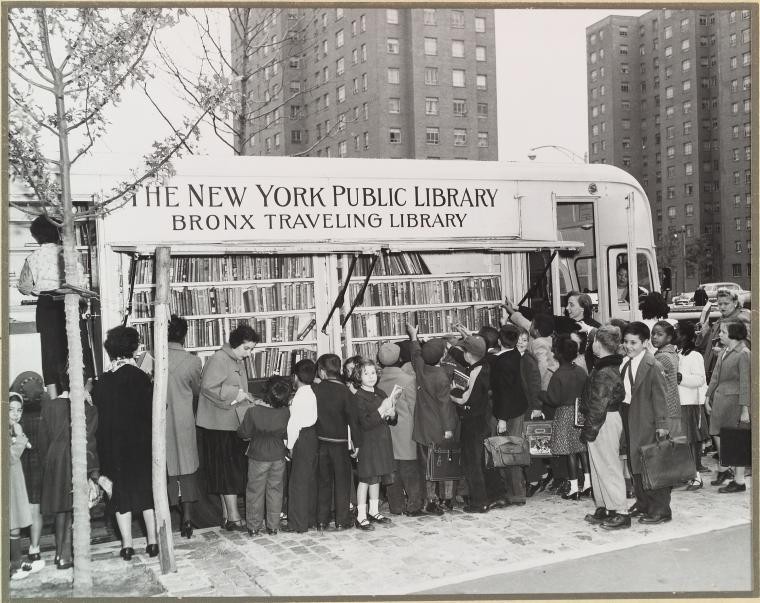 Black-and-White Photo of the New York Public Library Bronx Traveling Library, a bus with bookshelves built into the exterior sides. A crowd of people is gathered around the shelves checking out the books.