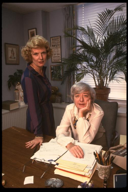 Two women, one sitting and another standing, are photographed behind a desk