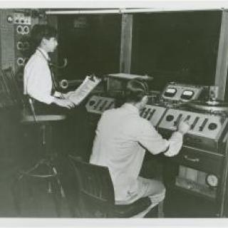 Black and white photo of two people using older sound recording technology.