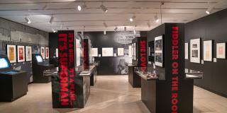 Black columns printed with the titles of Harold Prince directed musical productions in red printed on their side are shown in the main exhibition space. Posters and manuscript materials along with several kiosks and display cases line the room. 