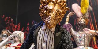 Figure wears an ornate gold mask of a lions face and an embroidered dress shirt and coat; mannequins in background wear fancy dress including headdress with several large feathers standing up.