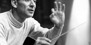 Leonard Bernstein is shown one hand raised palm facing the camera the other with conducting baton.  