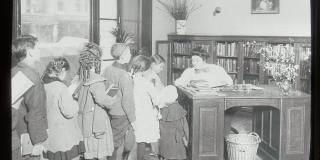Black and white photograph sitting at a desk with a long line of children dressed in 20th century fashion.