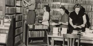 Black and white photograph of four women sorting through a library cart and stacks of books.