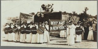 Black-and-white photograph of women from different states dressed all in white and parading for suffrage with banners reading "Government By Consent" and "Why Not in New York?"