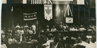 Black-and-white photograph of Carrie Chapman Catt opening the International Woman Suffrage Alliance Congress in 1913. Flags of the United States and the United Kingdom are draped behind her.