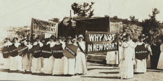 Historical black-and-white photograph of women from different states campaigning for suffrage and carrying a banner reading "WHY NOT IN NEW YORK?"