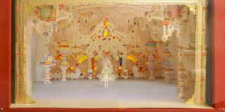 A photo of a set model with red fabric sides. The proscenium is made out of gold lace with pink, blue, and yellow jewels across the top. The interior of a castle with a throne is made out of white lace covered in colorful candy. A ballerina in a green tutu is standing the center.