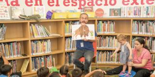 Photo of a man during a storytime event in a library, showing a picture book to a group of children sitting in a room full of bookshelves