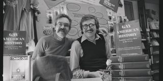 Two men, both with moustaches and glasses, standing at a desk with books saying "Gay American History" next to them 