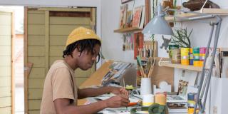 Photograph of illustrator Christian Robinson painting at a work table filled with paint and other art materials
