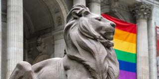 Photo of a marble lion statue in front of the Stephen A. Schwarzman Building, which features a large rainbow banner on the facade