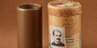 Two cylinders sitting within a brown background. The cylinder on the left is solid brown, and the one on the right is slightly lighter and larger with the image of a man's face on it