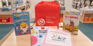 Photo of a children's summer book kit, which includes activity packets, two books, and a red drawstring backpack with the NYPL logo and white text that reads: New York Public Library