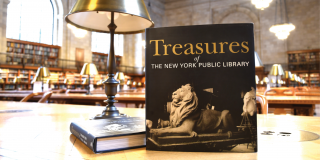 Photo of a book, Treasures of The New York Public Library, displayed on a table in the Rose Main Reading Room next to a gold lamp