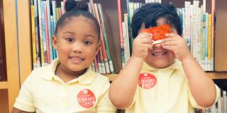 Photo of two children in yellow polo shirts with red stickers; one child is holding a library card in front of their eyes while smiling