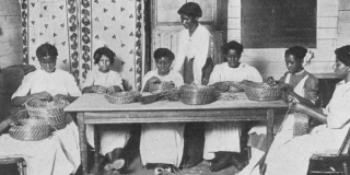 Historic black-and-white photo eight Black women sitting around a long table, wearing white dresses, and weaving baskets