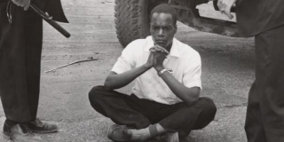 Historic photo of a Black man in a white shirt sitting cross-legged on the ground with his hands folded underneath his chin while two cops standing next to him, clutching clubs