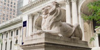 Statue of a marble lion outside of the Stephen A. Schwarzman Building, which is displaying a Treasures-themed dark blue banner.