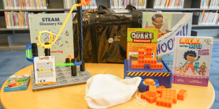 Photo of an NYPL Engineering Kit spread out on a table with items including: Roller Coaster Challenge puzzle, Survive the Quake game, and two books