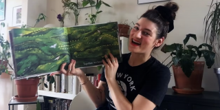 Video still of an NYPL librarian wearing an NYPL shirt and sitting in a room full of plants while holding up a picture book with illustrations of alligators.