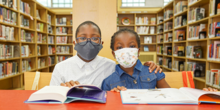 Two young Black children in face masks sit at a red table with two open books. 