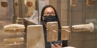 A woman in glasses and a black face mask holds her phone up while looking into a display case with ancient objects.