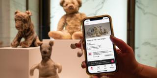 Photo of a hand holding an iPhone in front of the original Winnie-the-Pooh stuffed bear; the iPhone screen shows a Spanish-language audio guide. 