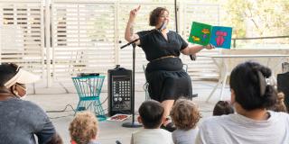 Photo of a librarian reading a picture book to a group of children and caregivers in an outdoor setting.