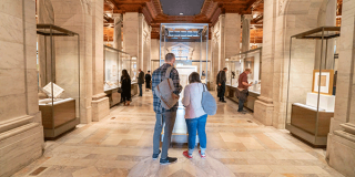 The Polonsky Exhibition in a large room with columns, and display cases featuring framed art work, books, and documents that people are leaning over to examine.