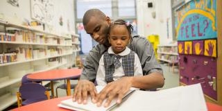 A man in a gray shirt and a small child in a white and plaid outfit read from a Braille book inside of a room with bookshelves and a colorful tactile wall display at Andrew Heiskell Braille and Talking Book Library. 