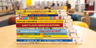 A stack of 13 children's library books sits atop a table in a well-lit room with colorful furniture. 