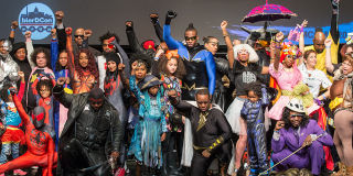 A large group of people cosplaying as comic book characters pose together on stage at the Schomburg Center. 