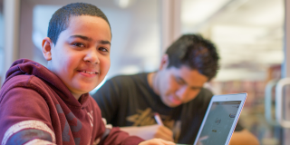 A smiling teen in a maroon hoodie smiles in the foreground next to a laptop; in the background, another teen in a black shirt is focused on writing. 