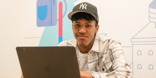 Photo of a teen in a Yankees hat and a button-down shirt sitting down with a laptop in front of a colorful mural. 