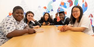 A diverse group of teens sits at a long wooden table, smiling at the camera; in the background is a colorful mural. 