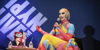 Sasha Velour sits on the LIVE from NYPL stage holding a microphone and wearing a brightly colored outfit. 