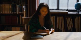 Sally Wen Mao, an Asian woman with long hair wearing a green-and-black dress, sits at a table in front of bookshelves, writing in a notebook