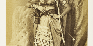 Portrait of a standing woman in ornate draped clothing and jewelry posing next to a plush chair.