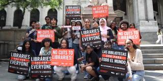 A group of teens outside the Stephen A. Schwarzman building holding orange and black signs with slogans including "Libraries are for Everyone" and "No Cuts to Libraries!"