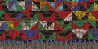 Beadwork in triangular and geometric patterns of many colors, attached to a threaded “belt” and decorated at bottom with a row of cowrie shells dangling from gathered threads