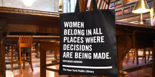 Black tote bag hanging from the back of a chair in the Rose Main Reading Room. White text on the bag reads: "Women belong in all places where decisions are being made," attributed to Ruth Bader Ginsburg.