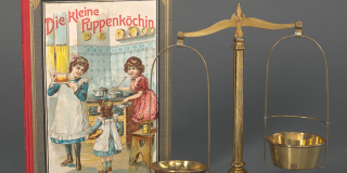 Photo of a historic book with an illustration of two women and a girl in a kitchen along with German text that reads: Die Kleine Puppenköchin; next to the book is a pair of brass scales and an assortment of small weights