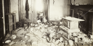 Historic black-and-white photo of a chaotic room inside of a New York City tenement with debris littered all over the floor, a wood-burning stove, an overturned chair, and a dusty jacket hung on a door; small naked child squats in a doorway in one of the few spaces where the floor is visible.