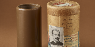 The wax cylinder is a dark brown tube, displayed upright. The containers echo the tubular shape of the cylinder. They are made of a lighter brown cardboard, with a round lid on the top. Some of them have handwritten notes written directly onto the cardboard, as well as paper labels and stickers. 