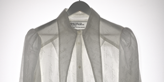 Long-sleeved white shirt with elongated triangular collar panels, backlit to highlight show the embroidered text that covers the fabric