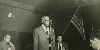 Black-and-white photograph showing William Patterson in a suit, standing at a microphone, with an individual seated in front of him and one either side, and an American flag in the background