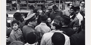 Black-and-white photograph showing a crowd of standing people surrounding a seated Dr. King, whose face is visible in the throng