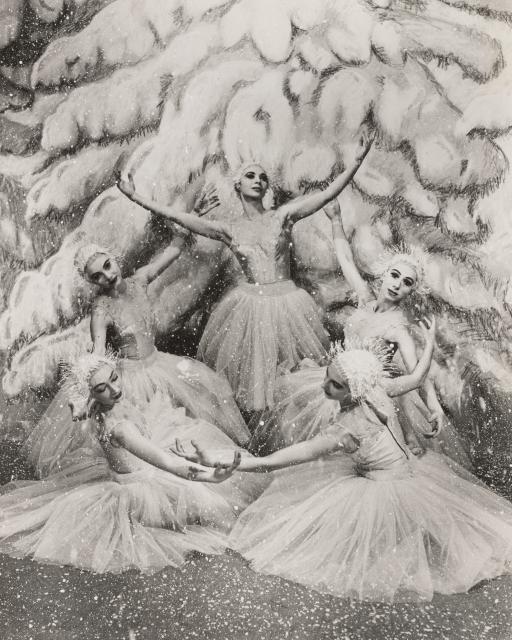 A black and whit photo of 5 dancers dressed as snowflakes forming a circle with their arms. A tree covered in snow is in the background.