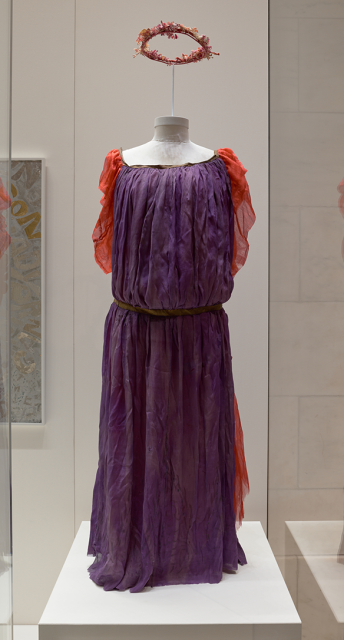 Costume of belted purple fabric with orange sleeves and a garland of flowers displayed above